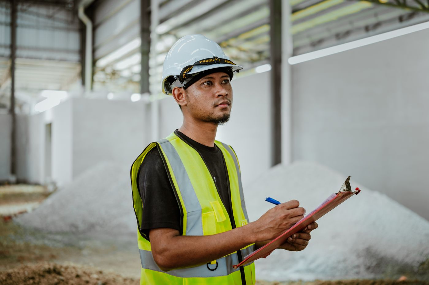 Profissional de capacete e colete conferindo uma prancheta no ambiente industrial
